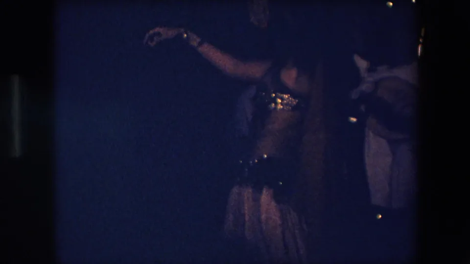 In 1971 Algeria home-movie footage, a dancer in a sparkling embellished costume raises an arm while musicians stand or sit nearby in very low blue light.