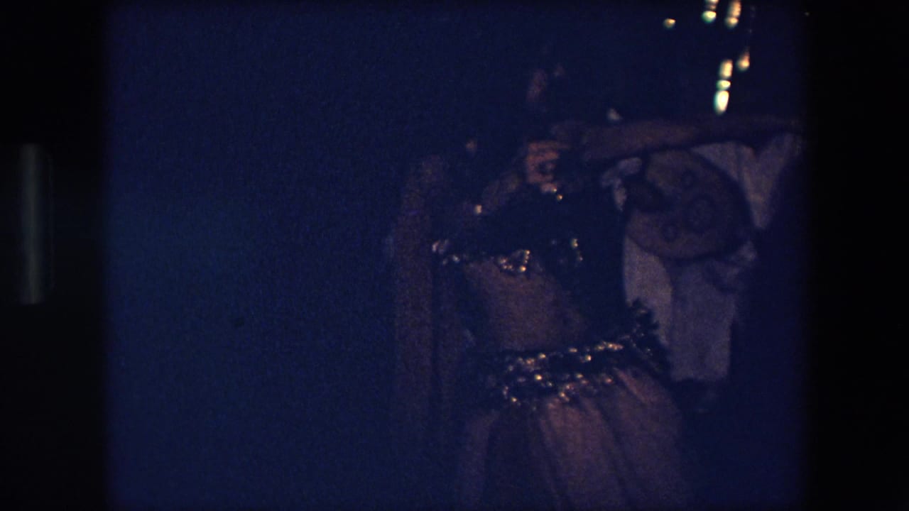 The dancer appears mid-movement in a dim blue-lit Algerian performance scene, with sequined costume details catching light and musicians partly visible behind her.
