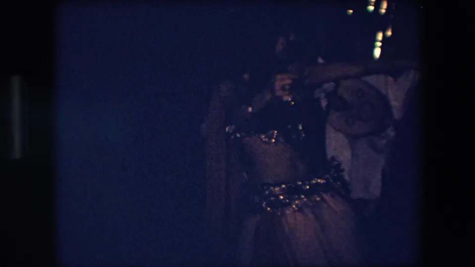 The dancer appears mid-movement in a dim blue-lit Algerian performance scene, with sequined costume details catching light and musicians partly visible behind her.