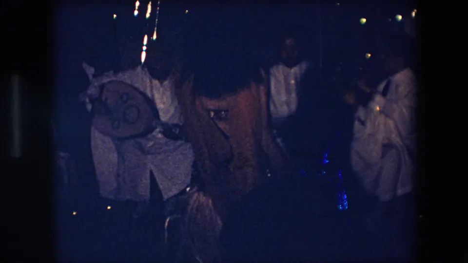 In the low-light 8mm frame, musicians in light traditional garments gather around percussion while the dancer is partly visible near the lower center of the scene.