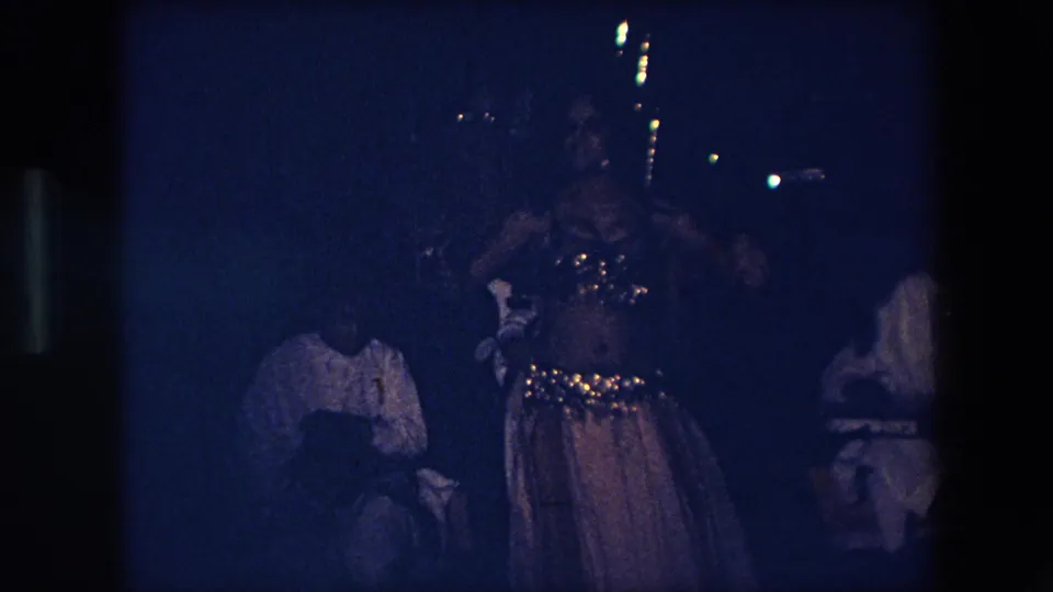 A dancer in an embellished costume stands near seated musicians in a dark blue-lit 1971 Algeria home-movie frame, with faces and instruments only partly legible.