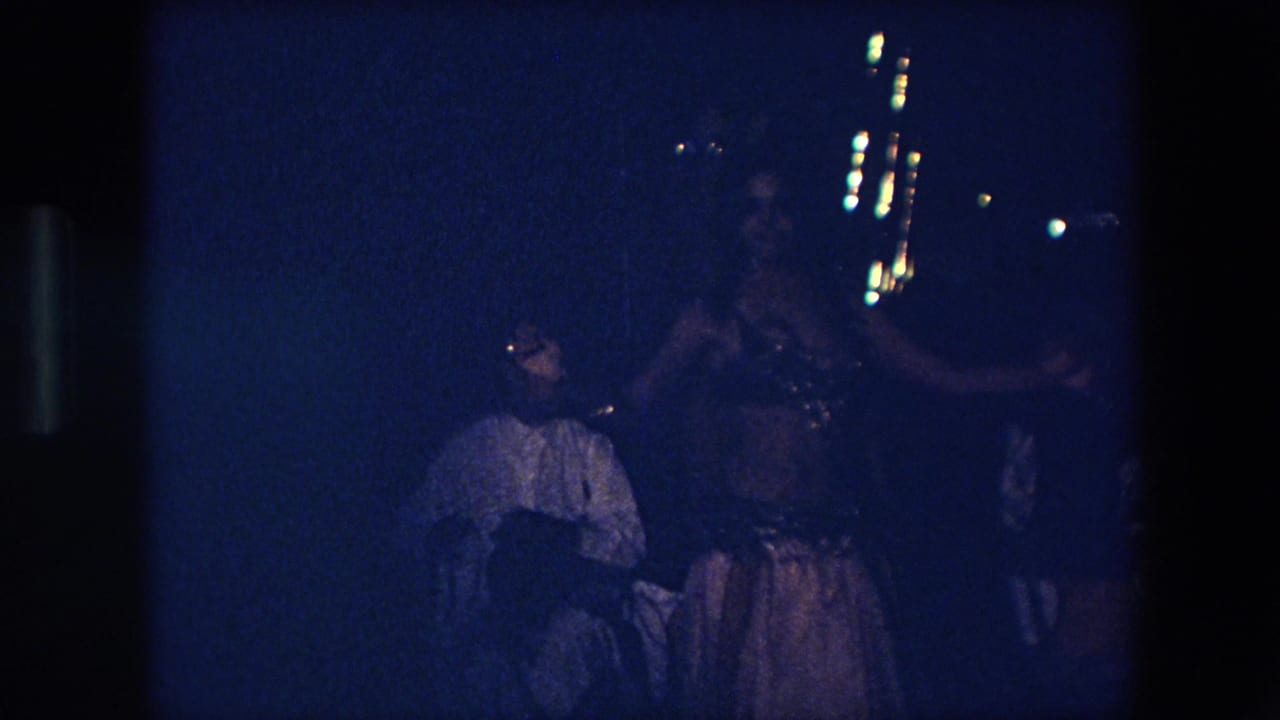 In Algeria in 1971, a female dancer appears in an ornate costume under low blue light while a seated audience member watches in the foreground.