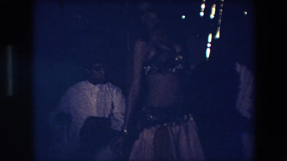 A 1971 Algeria home-movie frame shows the dancer near seated viewers, with her costume and hip belt partly visible through grain and low exposure.