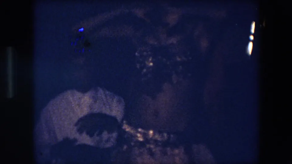 The 8mm frame moves closer to the dancer's torso and beaded costume while a seated person remains partially visible in the dim blue foreground.