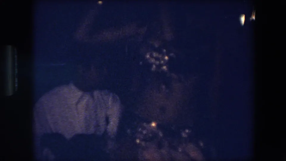 1971 Algeria 8mm frame showing a dark, blue-tinted interior with a person in a light-colored shirt and indistinct people nearby.