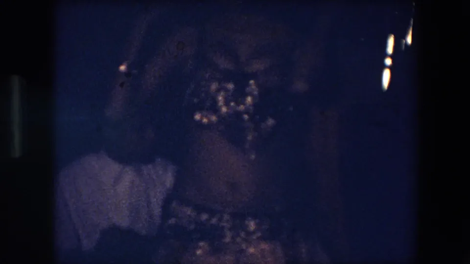 1971 Algeria home-movie frame with a shadowed person in a dim room and a partial figure wearing a light-colored shirt.