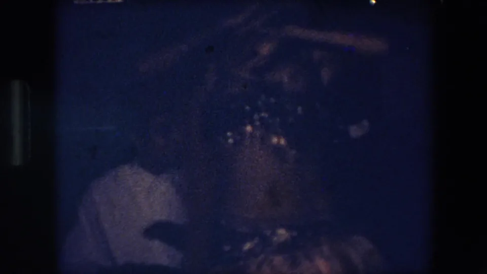 1971 Algeria home-movie frame showing a person in a light-colored shirt beside another shadowed figure in a dark blue-tinted room.