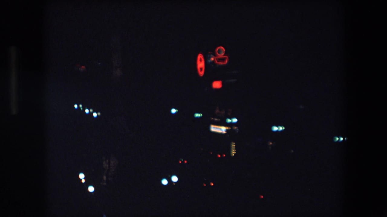 1971 Barcelona 8mm night frame with scattered city lights and a small red neon camera-shaped sign glowing against a dark background.