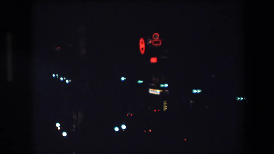 1971 Barcelona 8mm night frame with scattered city lights and a small red neon camera-shaped sign glowing against a dark background.