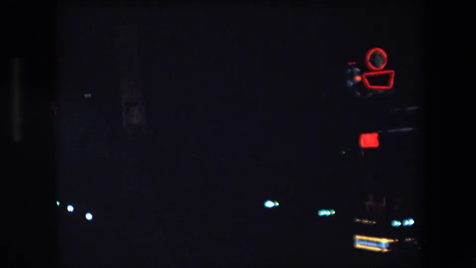 1971 Barcelona 8mm night view with a red neon camera-shaped sign near the upper right and sparse blue-white lights across the darkness.