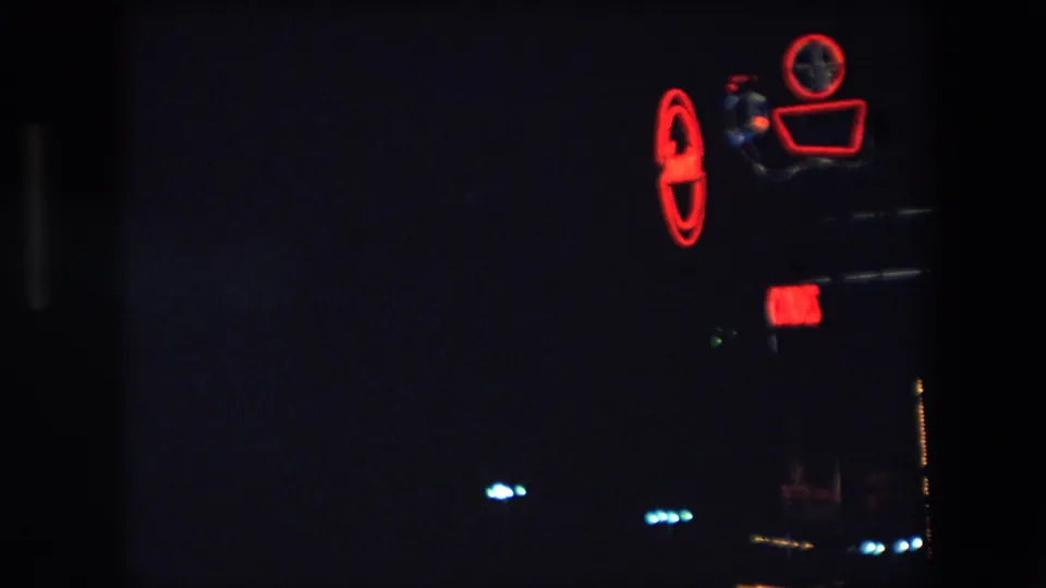 Closer 1971 Barcelona 8mm frame of a red neon camera-shaped sign glowing in a very dark urban night scene, with small illuminated details below.
