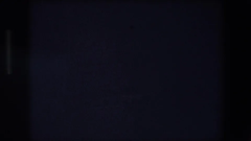 Underexposed 8mm frame from the Barcelona clip showing a dark panel-like surface, a broad vertical shadow, and barely visible light traces.