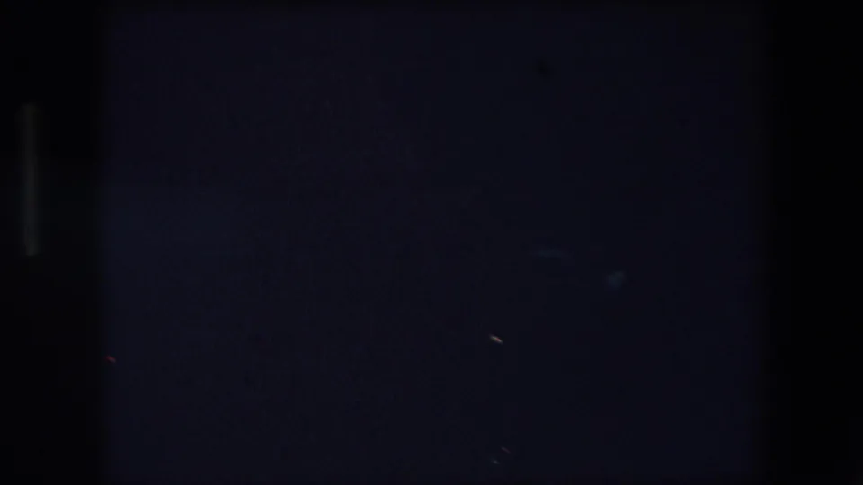 Dim 1971 Barcelona home-movie frame where small red and blue lights begin to appear against a largely black control-station or sign area.