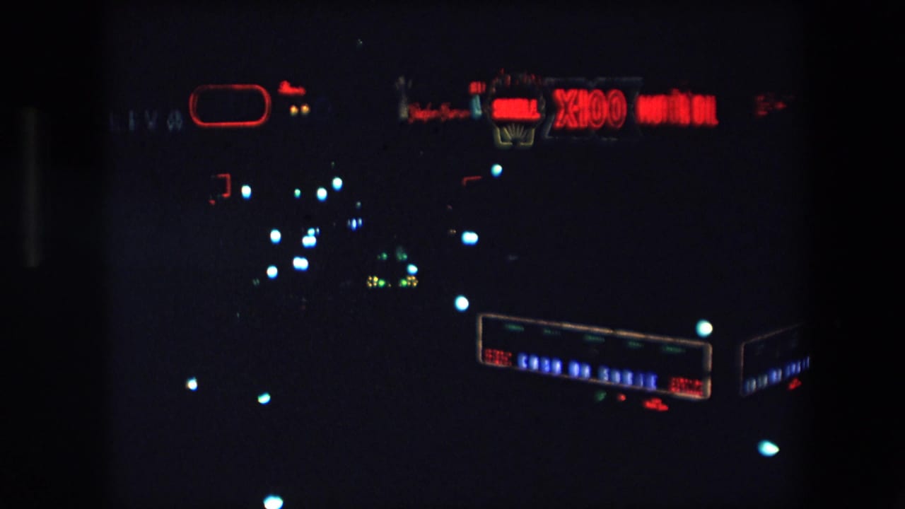 1971 Barcelona nighttime frame with a red Shell X100 Motor Oil sign, dark skyline, and soft blue-white points of city light.