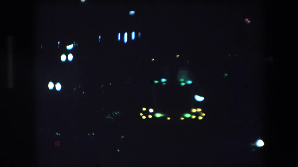 1971 Barcelona nighttime frame with out-of-focus white and green light clusters and faint colored points in deep shadow.