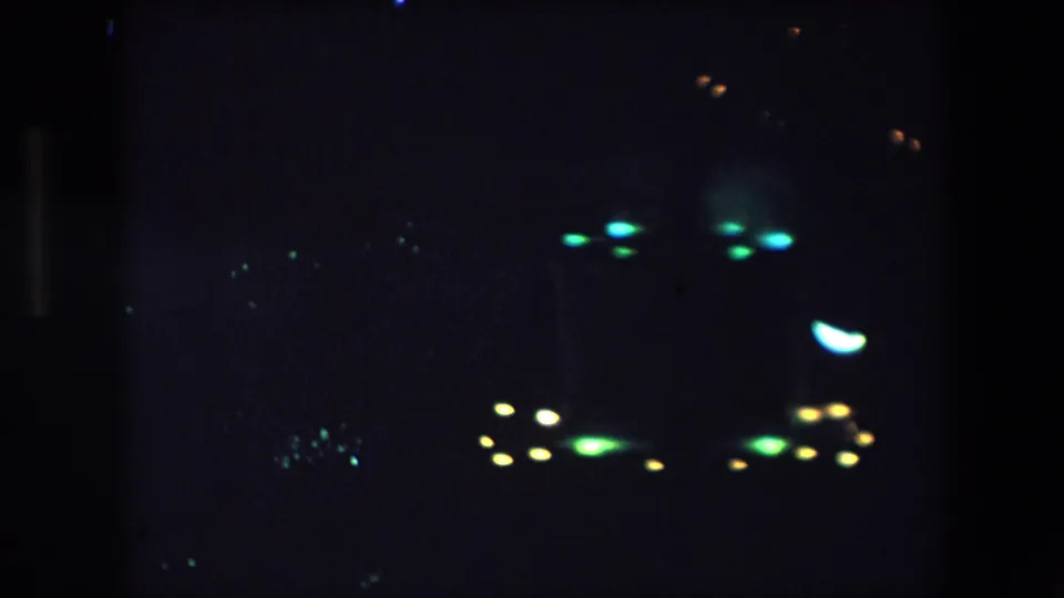 1971 Barcelona home-movie still with clustered green, yellow, and white lights floating in a dark underexposed night scene.