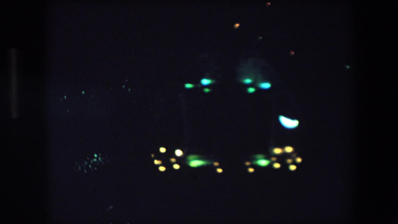 1971 Barcelona 8mm frame showing a dark night sky with distant clusters of green, yellow, and blue glowing lights and visible film grain.