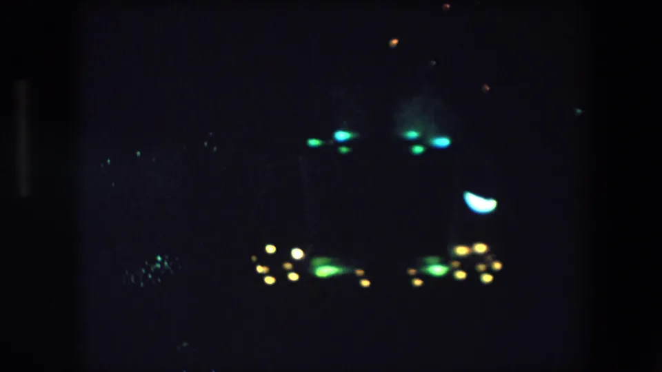 1971 Barcelona 8mm frame showing a dark night sky with distant clusters of green, yellow, and blue glowing lights and visible film grain.