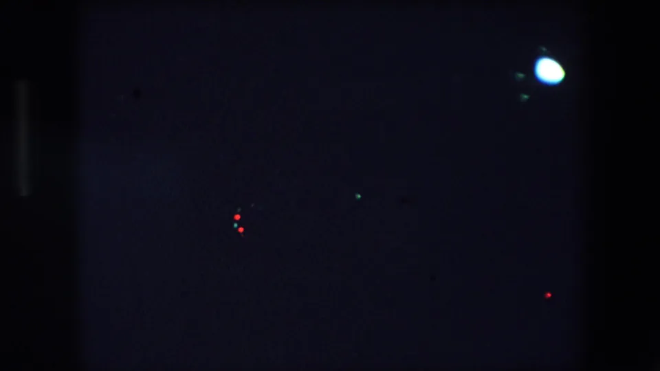 Nighttime 8mm frame from Barcelona with a bright circular glare, small red light points, and surrounding darkness from a moving urban view.