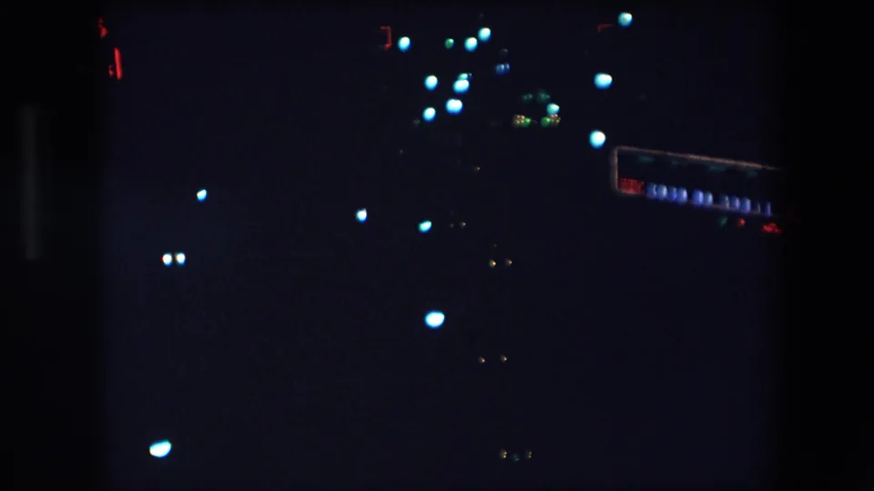 1971 Barcelona nighttime frame with clustered blue-white lights, tiny red points, and a faint rectangular illuminated shape that may be signage or a lit vehicle side.