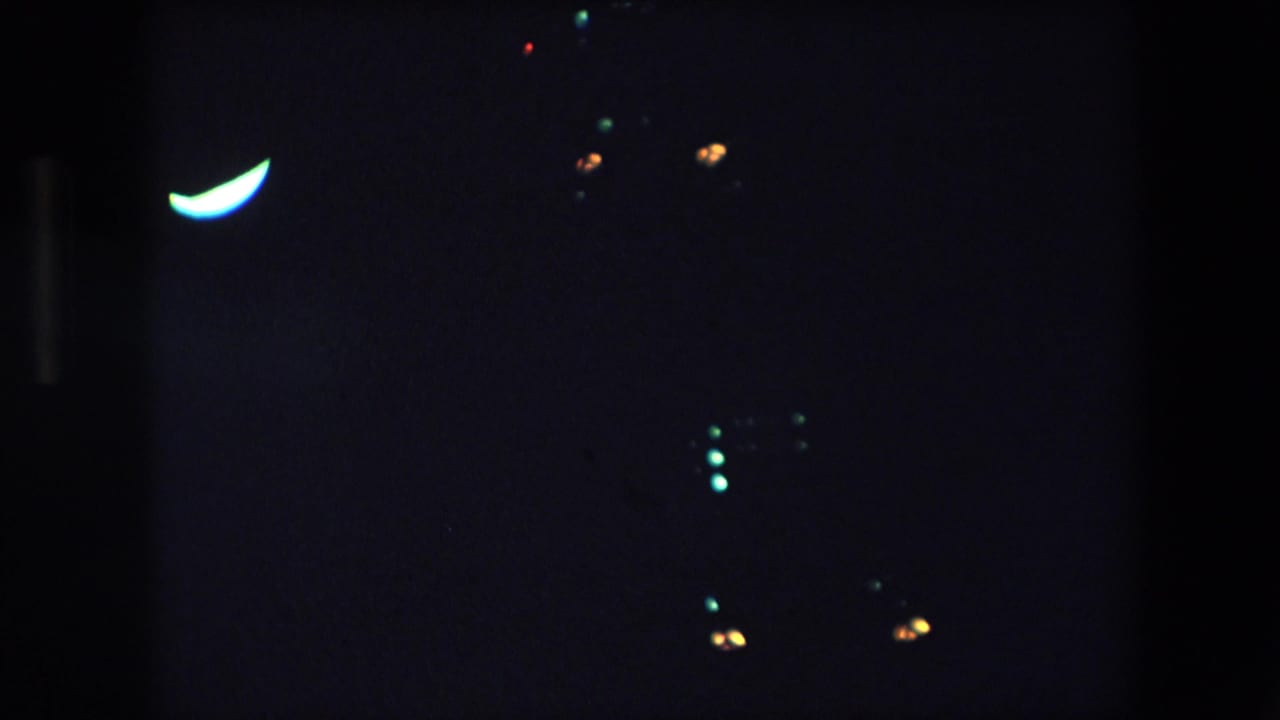1971 Barcelona 8mm frame showing a dark night view with a bright crescent-shaped light at upper left and scattered green and orange points in the surrounding darkness.