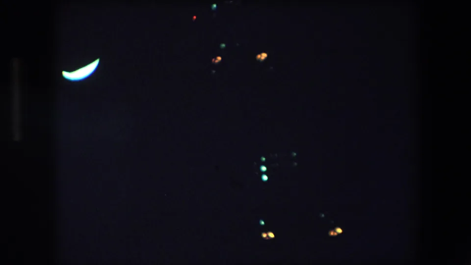 1971 Barcelona 8mm frame showing a dark night view with a bright crescent-shaped light at upper left and scattered green and orange points in the surrounding darkness.