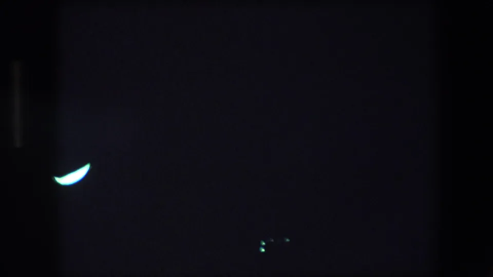 1971 Barcelona 8mm frame with a luminous crescent-shaped light against a nearly black field, with faint vertical dark bands and minimal visible surroundings.