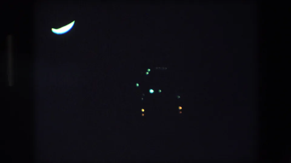 1971 Barcelona 8mm frame showing a crescent-like light at left and a loose cluster of small green, orange, and yellow lights lower in the dark scene.
