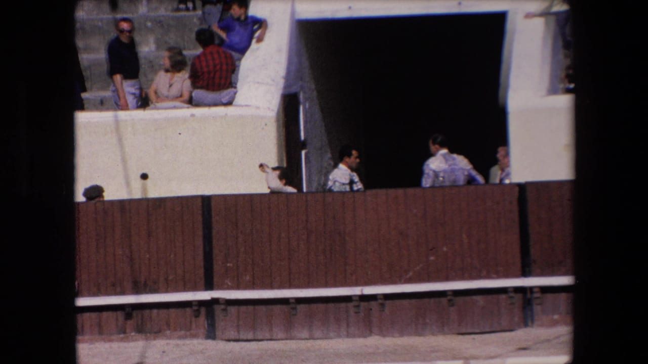 Spectators and attendants appear around a Barcelona bullring passageway, with a wooden barrier separating them from the arena floor.