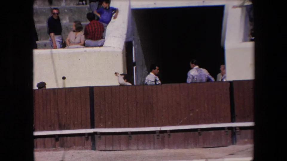 Spectators and attendants appear around a Barcelona bullring passageway, with a wooden barrier separating them from the arena floor.