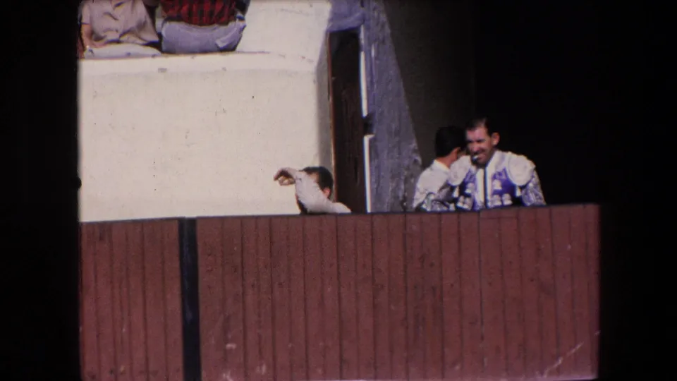 A matador in a decorated suit smiles behind the wooden bullring barrier as another matador stands near the dark entrance.