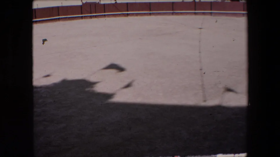 Barcelona 1971 8mm frame showing an empty sandy circular arena from a high viewpoint, with a red-and-white perimeter wall, dark shadow across the foreground, and a vertical obstruction at the side.