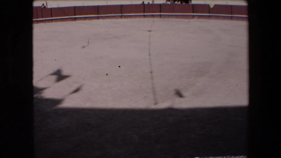 Barcelona 1971 8mm frame of the circular arena surface and red-and-white wall, partially divided by a dark vertical obstruction with shadowed foreground sand.