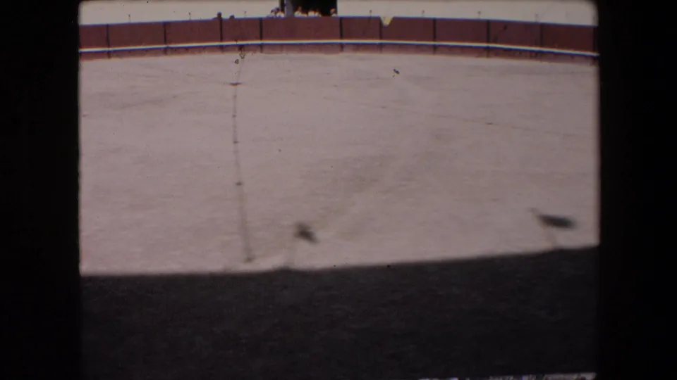 Barcelona 1971 8mm frame showing the empty sandy arena and curved red-and-white wall, with small distant spectators just visible above the perimeter near the upper edge.