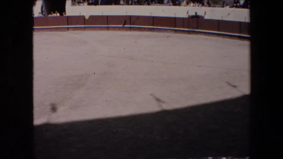 Barcelona 1971 8mm frame of the empty circular arena from above, showing the curved red-and-white perimeter wall, faint sand markings, foreground shadow, and tiny spectators at the upper rim.