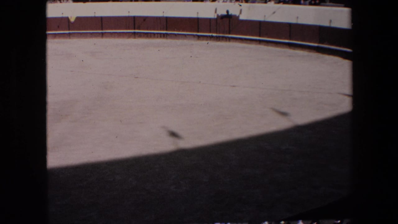 1971 Barcelona home-movie frame showing an empty bullfighting arena floor in bright sun, with a brown wooden fence and distant spectators behind white barriers.
