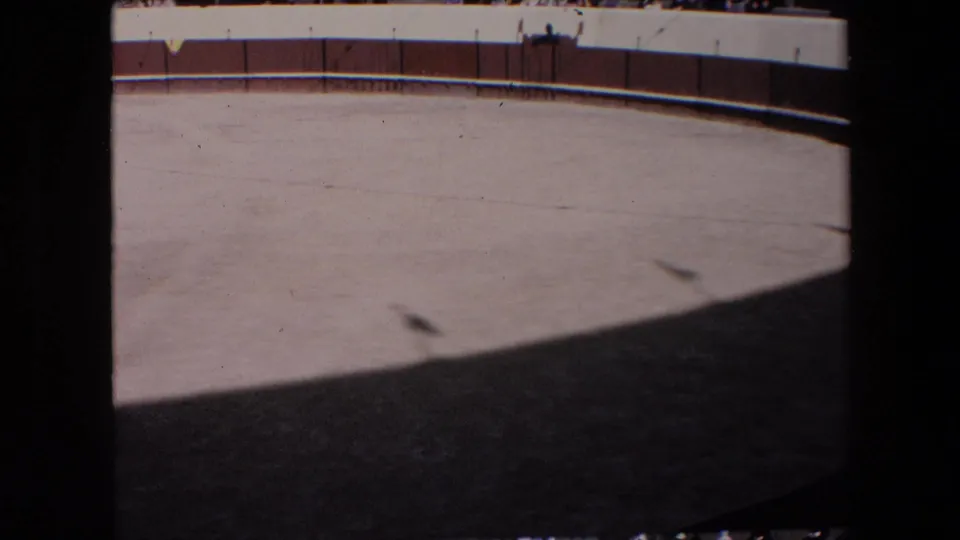 1971 Barcelona home-movie frame showing an empty bullfighting arena floor in bright sun, with a brown wooden fence and distant spectators behind white barriers.