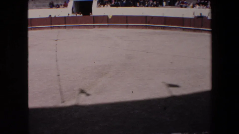 A sunlit 1971 Barcelona arena view shows the empty sandy ring, a central marking line, a wooden barrier, and distant spectators seated or standing behind the wall.