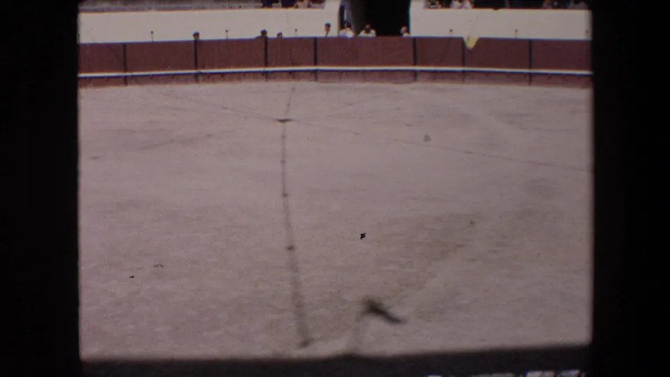 Wide 8mm frame from the Barcelona bullfighting arena showing a mostly empty sandy floor, central line, far wooden fence, white wall, and small spectators near the top edge.