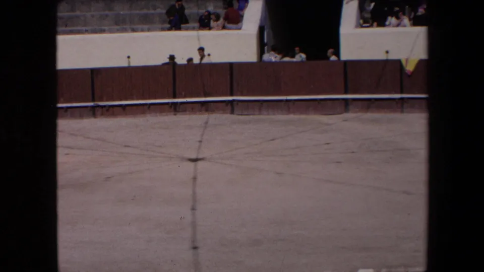 The final 1971 Barcelona frame holds closer on the far side of the bullfighting arena, with spectators above a brown wooden fence and an empty marked dirt floor below.
