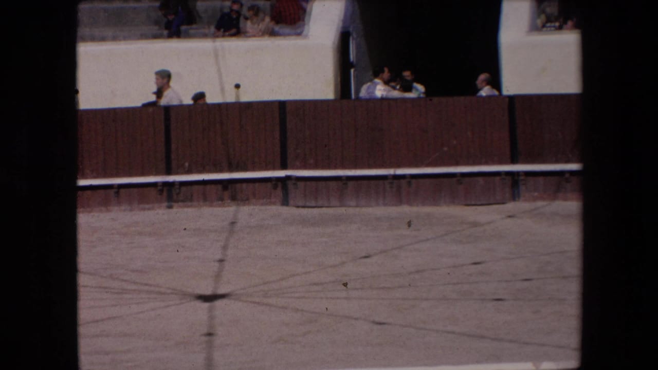 Barcelona 1971 home-movie frame showing a bullfighting arena edge, wooden barrier, concrete ring floor, and spectators seated above.