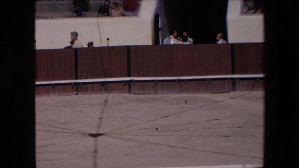 Barcelona 1971 home-movie frame showing a bullfighting arena edge, wooden barrier, concrete ring floor, and spectators seated above.