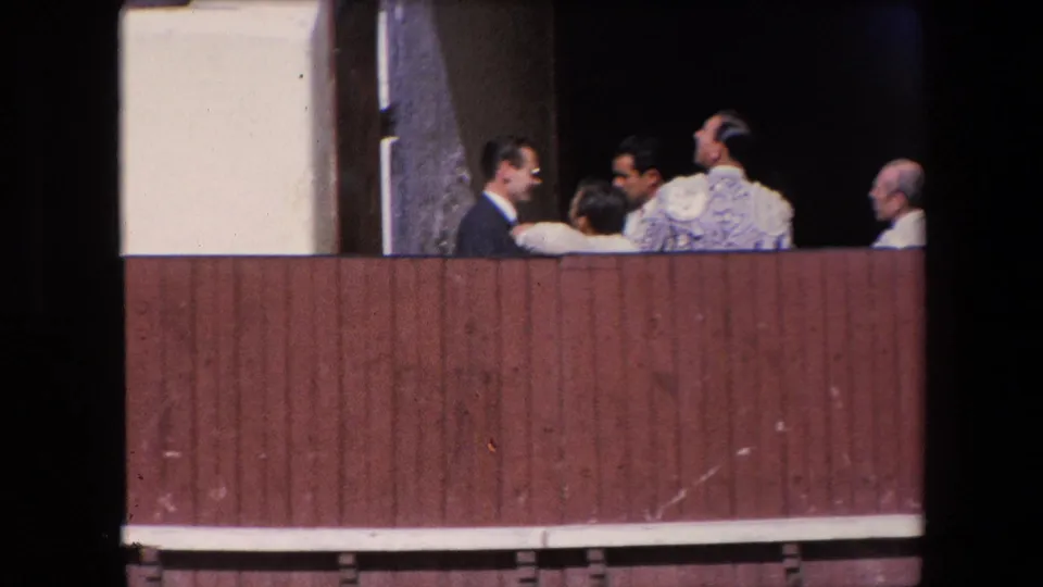 Barcelona 1971 home-movie frame showing suited men and a bullfighter or assistant in an ornate jacket gathered behind the wooden bullring barrier.