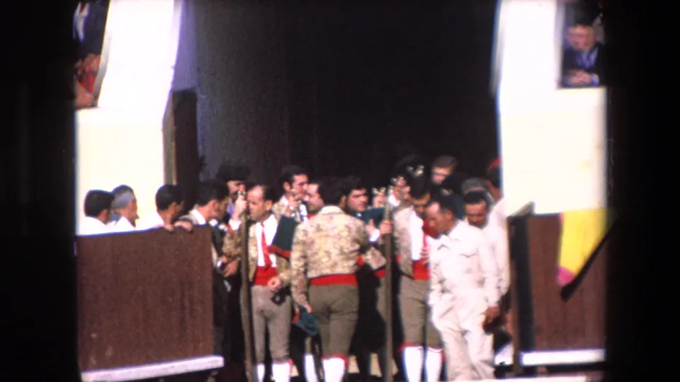 Barcelona 1971 frame showing bullfighters or assistants in decorated jackets and red sashes, a white-clad arena worker, and other men clustered beside the wooden arena barrier.