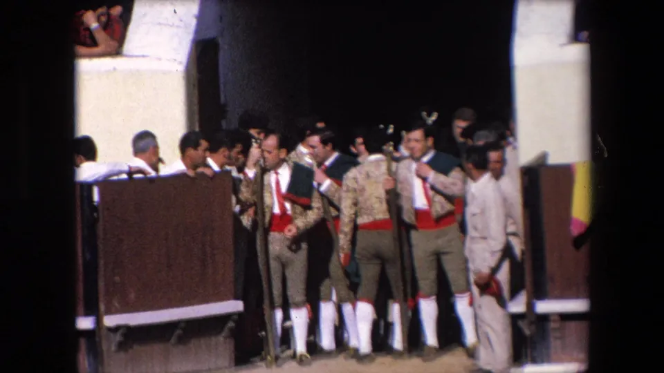 In supplied 1971 Barcelona footage, matadors in embroidered jackets, red sashes, and white socks stand with staffs near an open bullring gate while spectators watch.