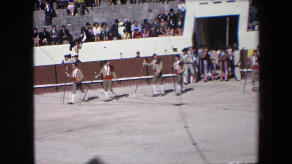 In the supplied Barcelona 1971 footage, matadors carrying staffs walk in formation across the sunlit sandy ring with spectators watching from tiered seating.