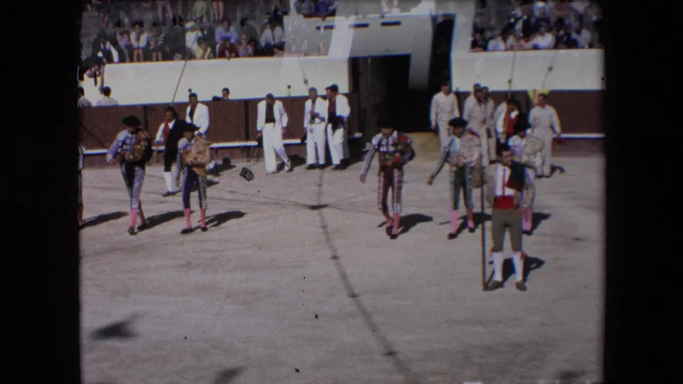 The 1971 Barcelona home-movie frame shows bullfighting participants in ornate dress, attendants in lighter clothing, and spectators surrounding the sunlit arena.
