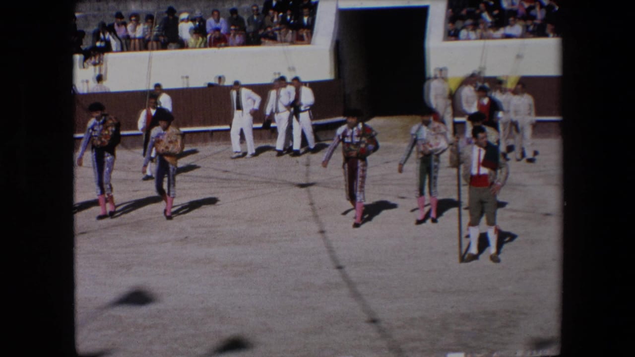 In 1971 Barcelona, bullfighters in ornate costumes and black monteras cross the arena floor while white-clad attendants and spectators are visible behind them.