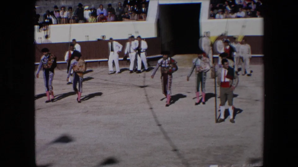 In 1971 Barcelona, bullfighters in ornate costumes and black monteras cross the arena floor while white-clad attendants and spectators are visible behind them.