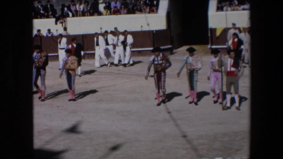 Bullfighters in colorful ceremonial dress walk in formation across a Barcelona bullring as spectators watch from the shaded and sunlit seating areas.